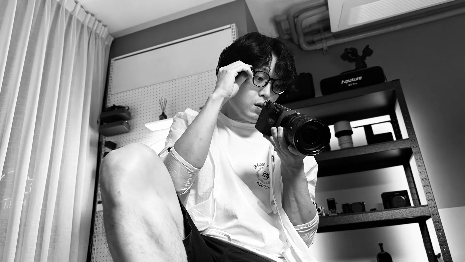 장비 선반 앞에 앉아 카메라를 들고 안경을 만지는 흑백 사진