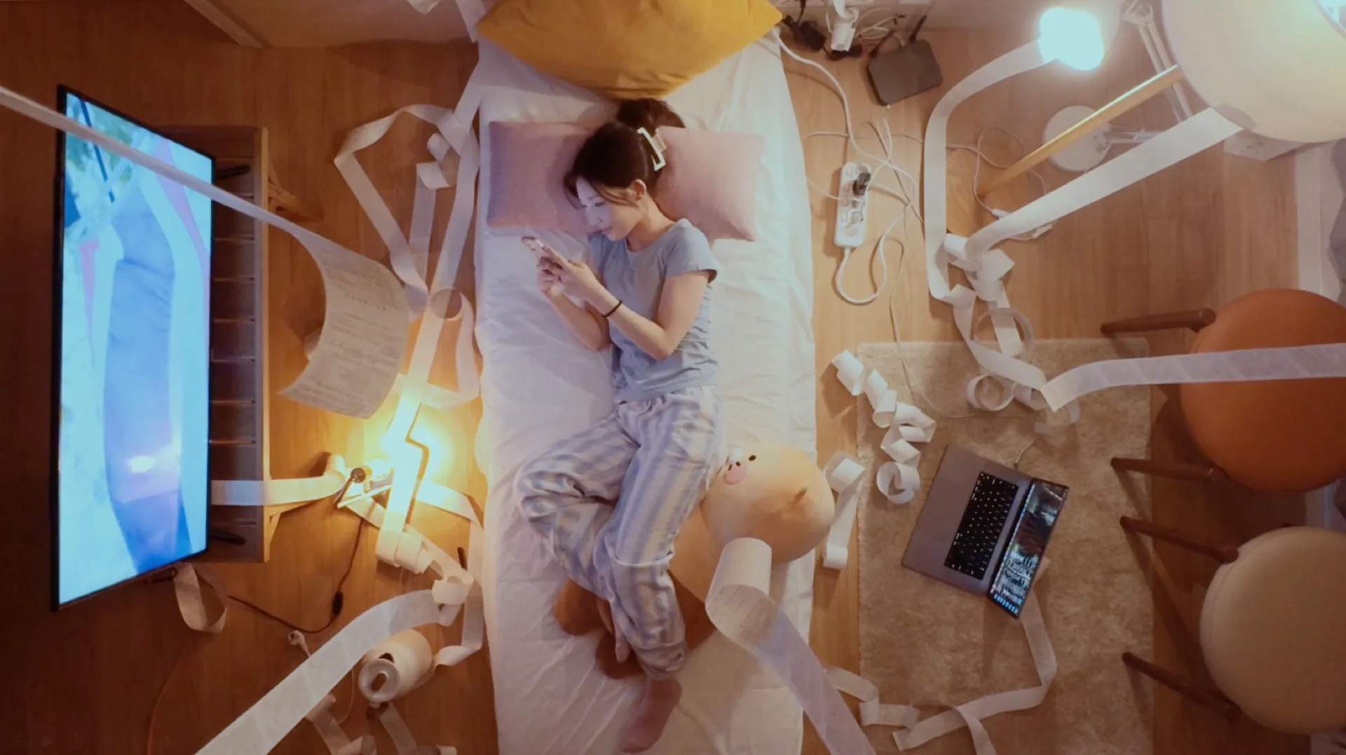 High-angle view of the whole room. Receipts pour from the TV, the lamp, the power strip and the laptop, and in the middle of it she lies on the bed looking at her phone
